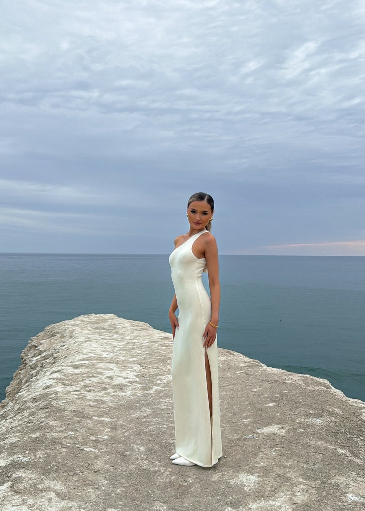 Woman in a white dress standing on a cliff overlooking the ocean. maia the label capri maxi dress