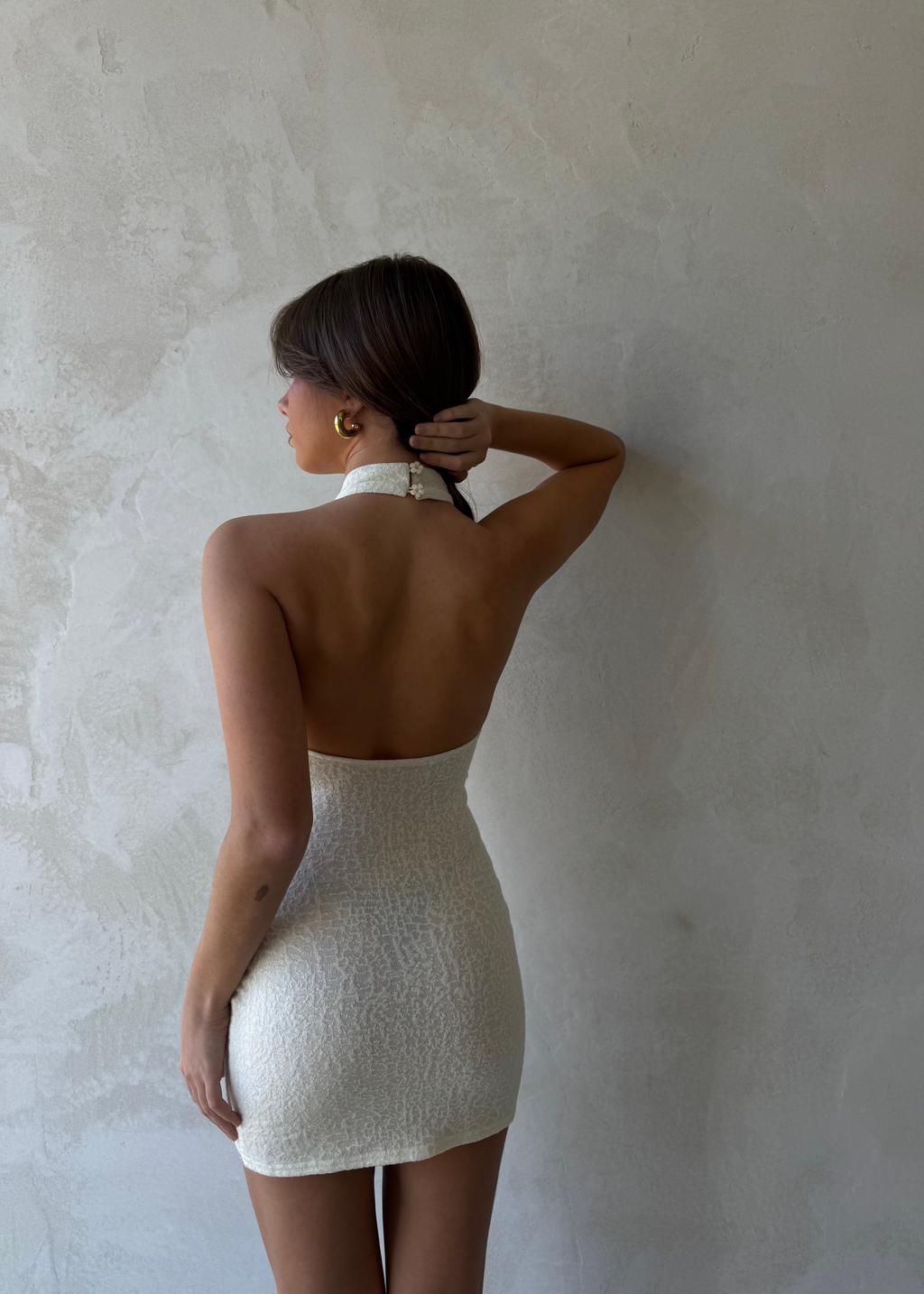 Back view of a woman wearing a dreamy meringue Dazie Mini Halter Dress with a high halter neck, low back, daisy button details, and a fitted wool blend fabric silhouette.
