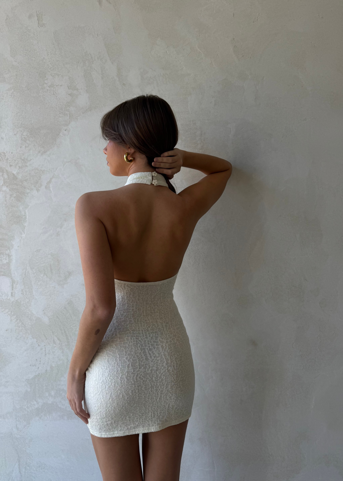 Back view of a woman wearing a dreamy meringue Dazie Mini Halter Dress with a high halter neck, low back, daisy button details, and a fitted wool blend fabric silhouette.