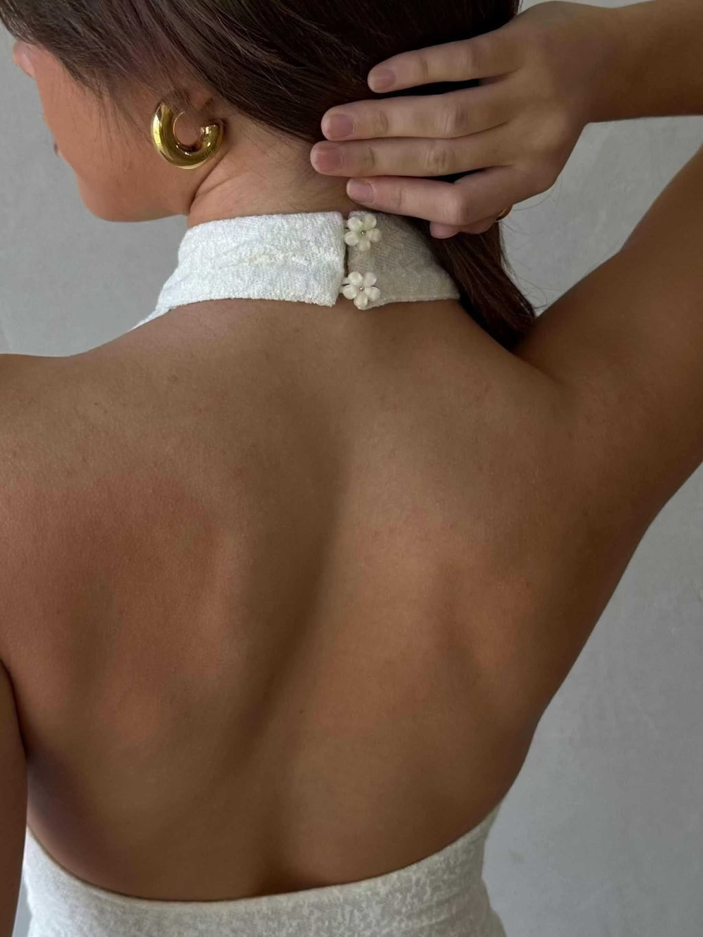 Back view of a woman wearing a white Dazie Mini Halter Dress with a high halter neck and daisy button details on the neck closure.
