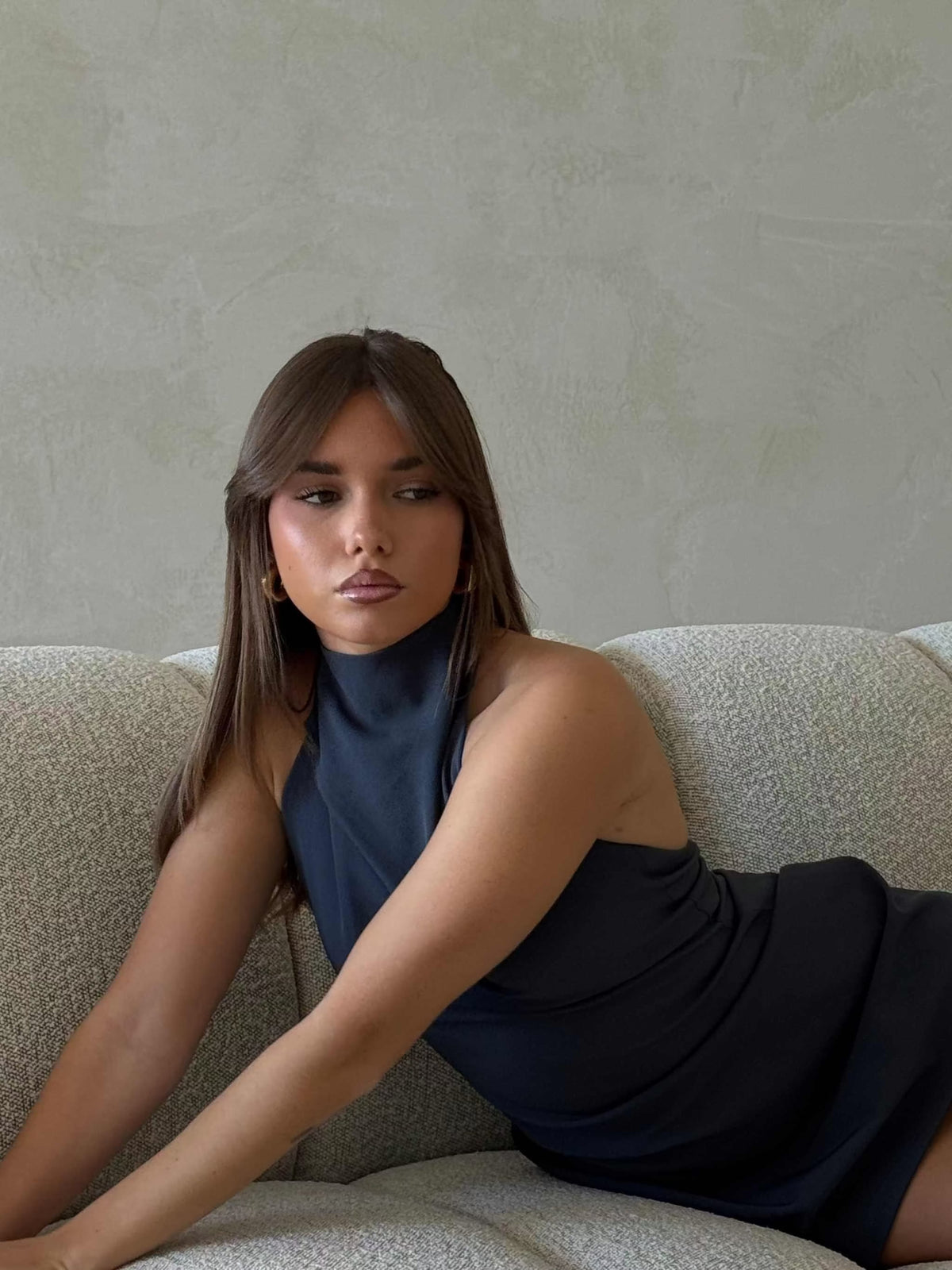 Woman wearing deep ocean blue Xara Mini Halter Dress with high halter neck and low back, sitting on a sofa