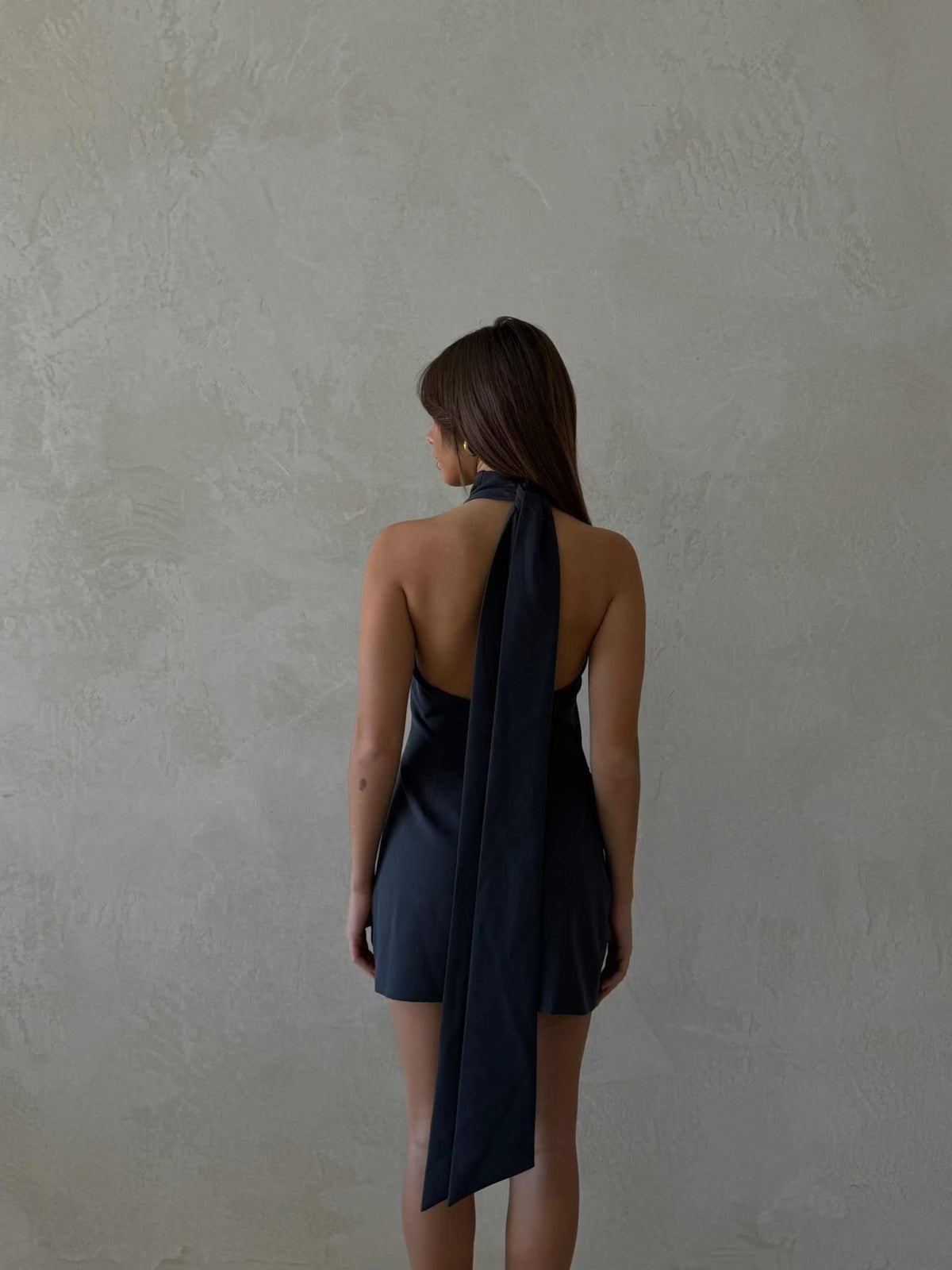 Back view of a woman wearing a deep ocean blue Xara Mini Halter Dress with a high halter neck and long flowing tie detail, featuring a low back and structured fabric.
