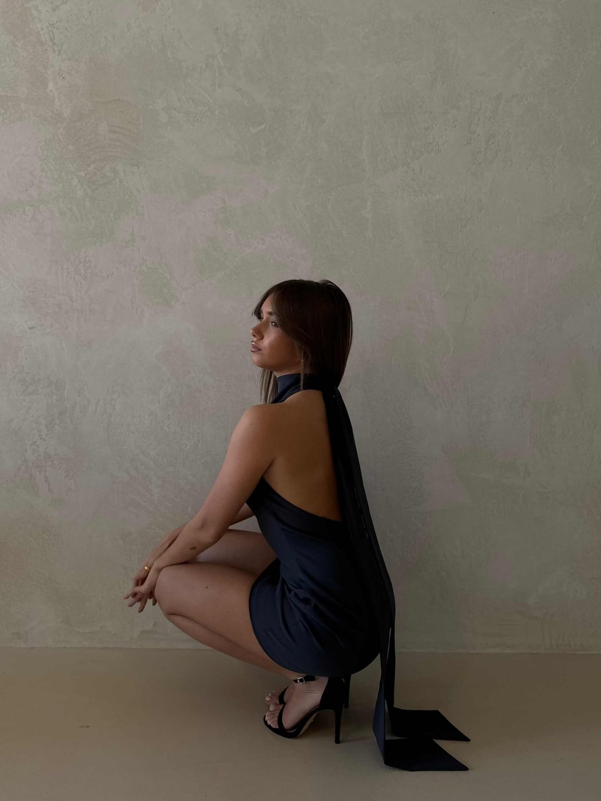 Woman wearing a deep ocean blue Xara Mini Halter Dress with a high halter neck, low back, and snatched waist, posing against a muted wall background.