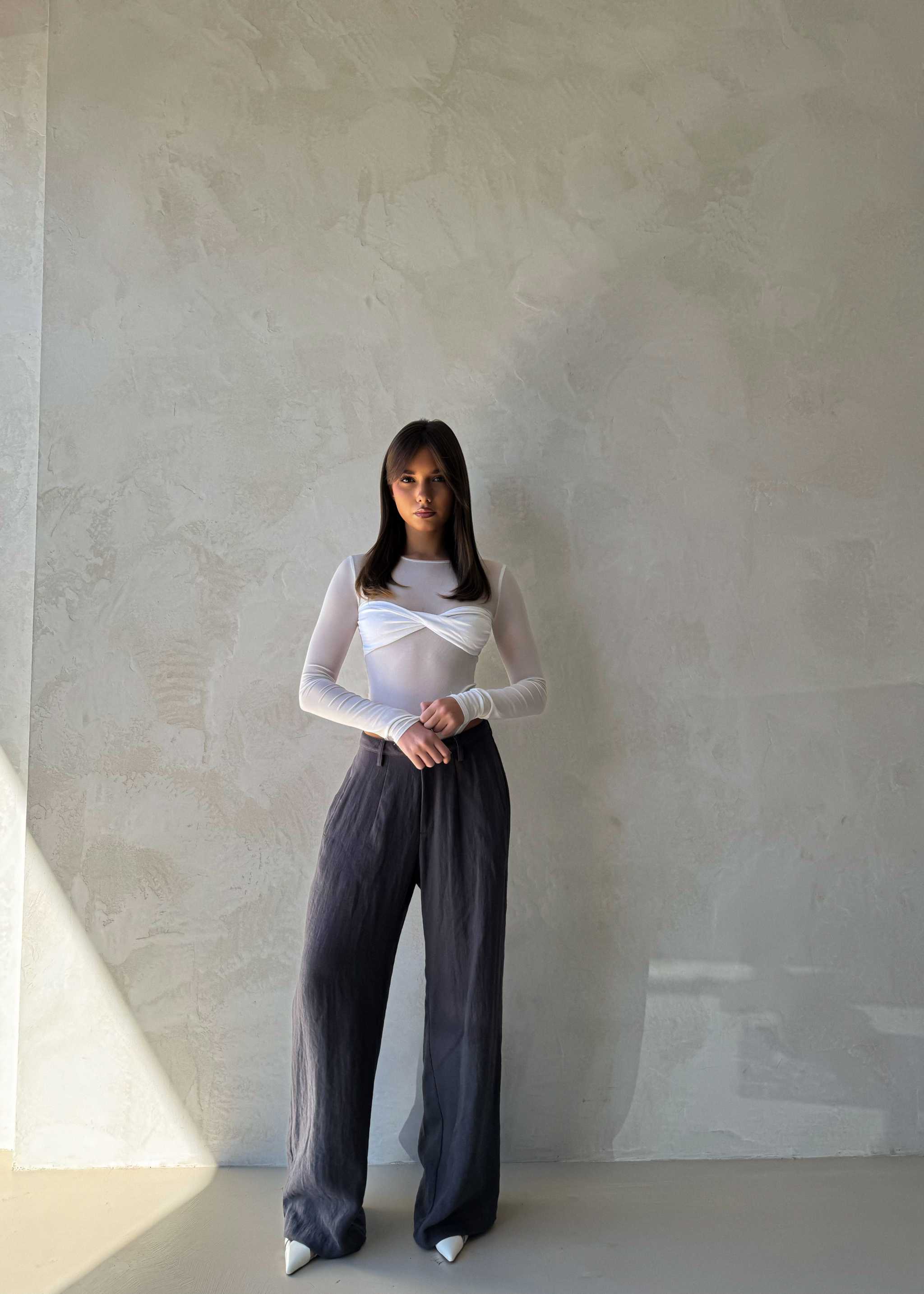 Arian Linen Pant Woman wearing a white long-sleeve top and gray wide-leg pants standing against a plain wall.