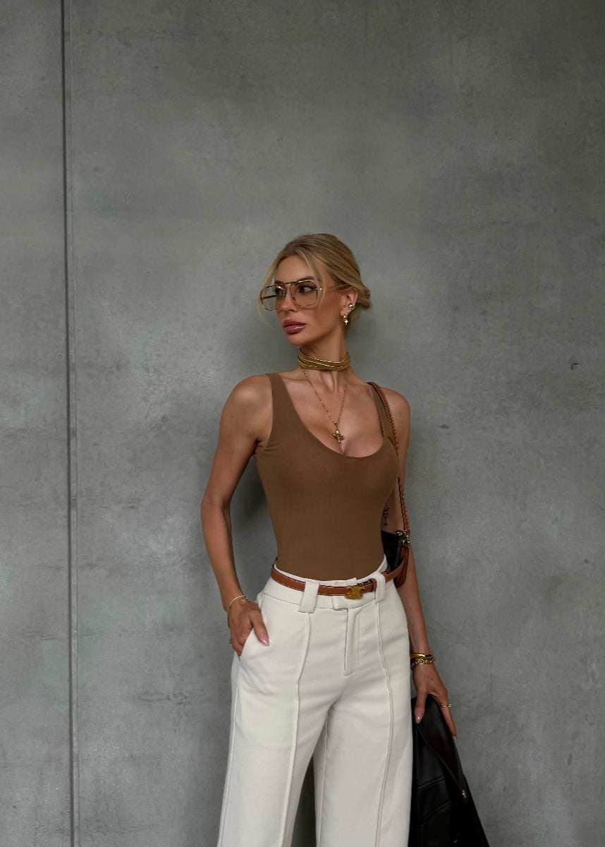 woman wearing rich chocolate Malu mesh bodysuit with deep neckline and white pants against grey wall